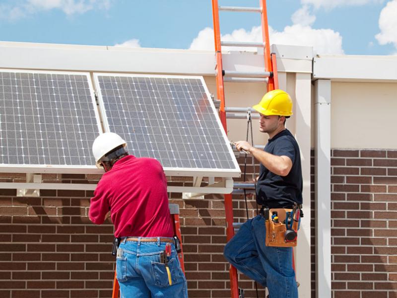 Two workers on ladders install solar panels on building